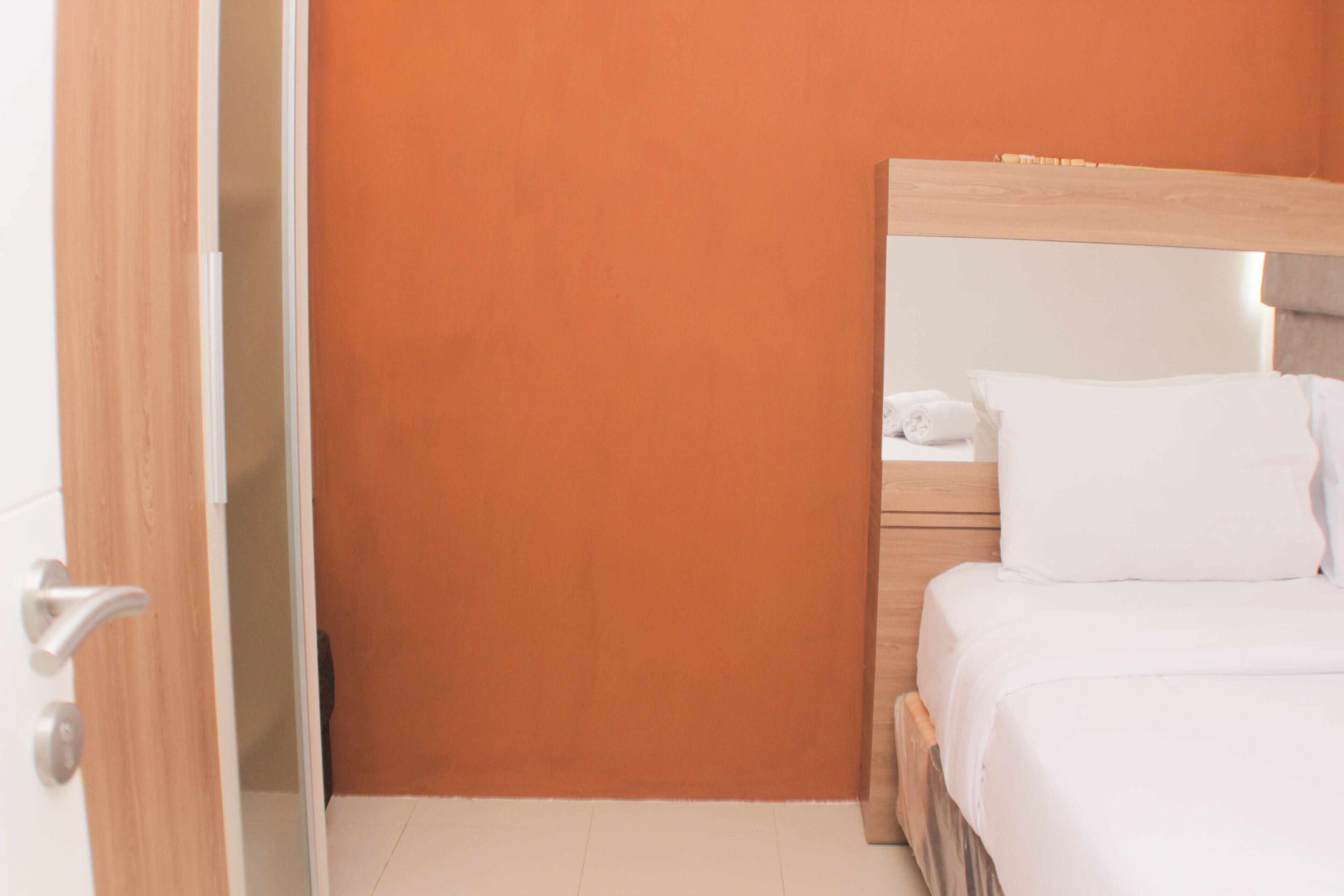 A hotel room with a wooden door, an orange wall, a wooden bed frame, and a white bedspread.
