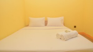 Un lit simple avec du linge de lit blanc et des oreillers, une serviette blanche roulée posée sur le lit, et un interrupteur sur le mur.