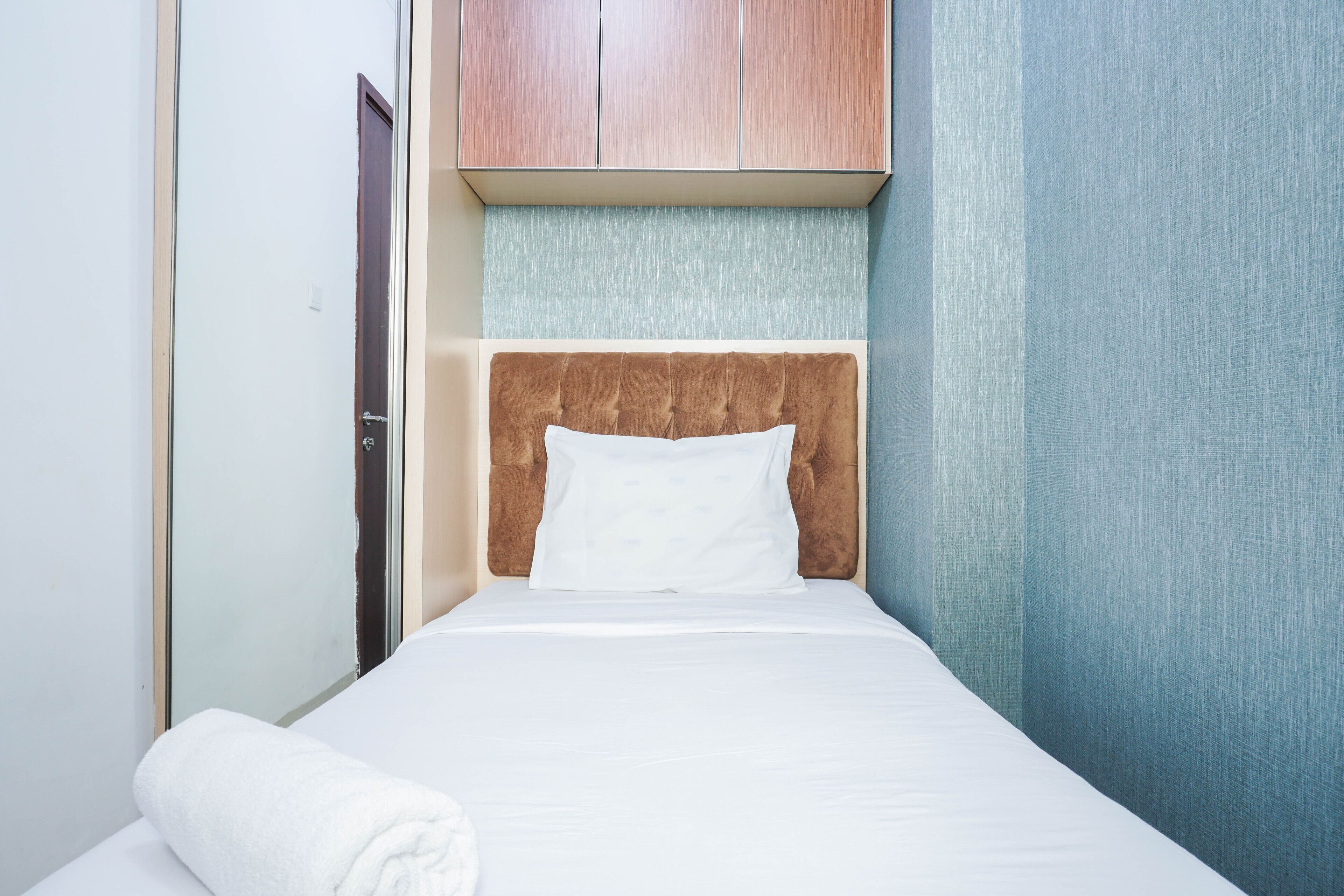 A single bed with a headboard, a wooden wardrobe above, and a towel on the floor.