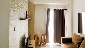 A living room with a brick wall, a flat-screen TV, two wooden stools, a brown sofa, and a window with brown curtains. - Graceful And Cozy 2Br At Signature Park Grande Apartment (Jakarta)