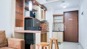 A compact kitchen with a microwave, refrigerator, and wooden cabinets. There are two stools and a sofa in the living area. - Graceful And Cozy 2Br At Signature Park Grande Apartment (Jakarta)