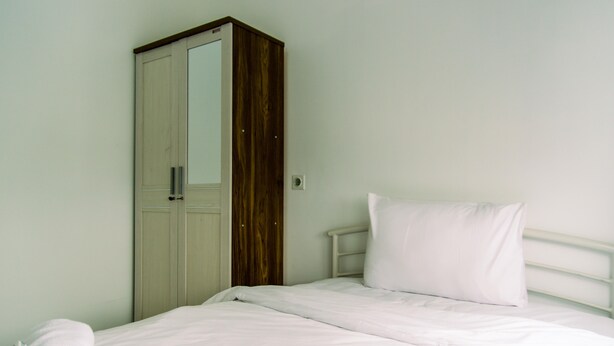 Una cama individual con ropa blanca, un armario de madera y un cabecero de metal blanco.