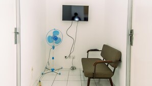 Una habitación con una televisión montada en la pared, un ventilador de pie y una silla.