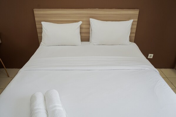 A neatly made bed with white linens and two pillows, a wooden headboard, and a plain brown wall.