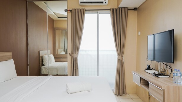 A hotel room with a large bed, a desk with a flat-screen TV, and a view of the city through the window. - Modern And Relax Studio Room At Serpong Greenview Apartment (South Tangerang)