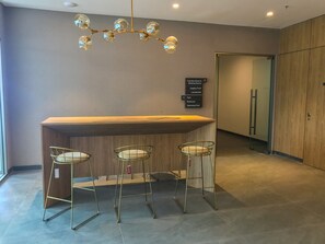 Un área de recepción moderna con un mostrador de madera, tres taburetes metálicos, un cartel indicador y una puerta de cristal que da a un pasillo.