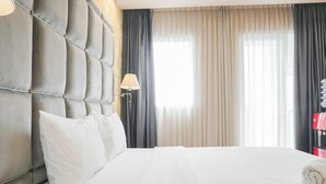 Una camera d'albergo con una testata imbottita, un letto con lenzuola bianche, una lampada e una finestra con tende.