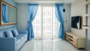 A modern living room with a blue sofa, a TV, and a sliding glass door with blue curtains. - Spacious And Nice 2Br At Casa De Parco Apartment (Pagedangan)