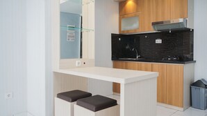 A modern kitchen with a white countertop, black backsplash, and wooden cabinets. There are two stools at the counter and a trash bin to the side. - Spacious And Nice 2Br At Casa De Parco Apartment (Pagedangan)