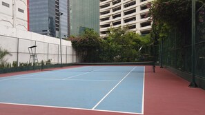 An outdoor tennis court with a red and blue surface, surrounded by a chain-link fence and greenery.