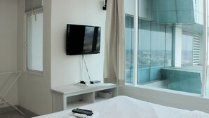 A hotel room with a bed, a TV mounted on the wall, a window with a view, and a white console table. - Warm And Cozy Studio At Grand Kamala Lagoon Apartment (Bekasi)