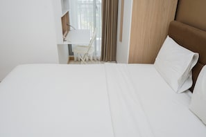 A hotel room with a neatly made bed, a wooden headboard, a small desk with a chair, and a window with curtains.