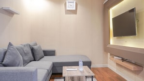 Un salon moderne avec un canapé d’angle gris, une petite table basse en bois sur laquelle se trouvent une télécommande et une bouteille d’eau, un téléviseur fixé au mur et une horloge.