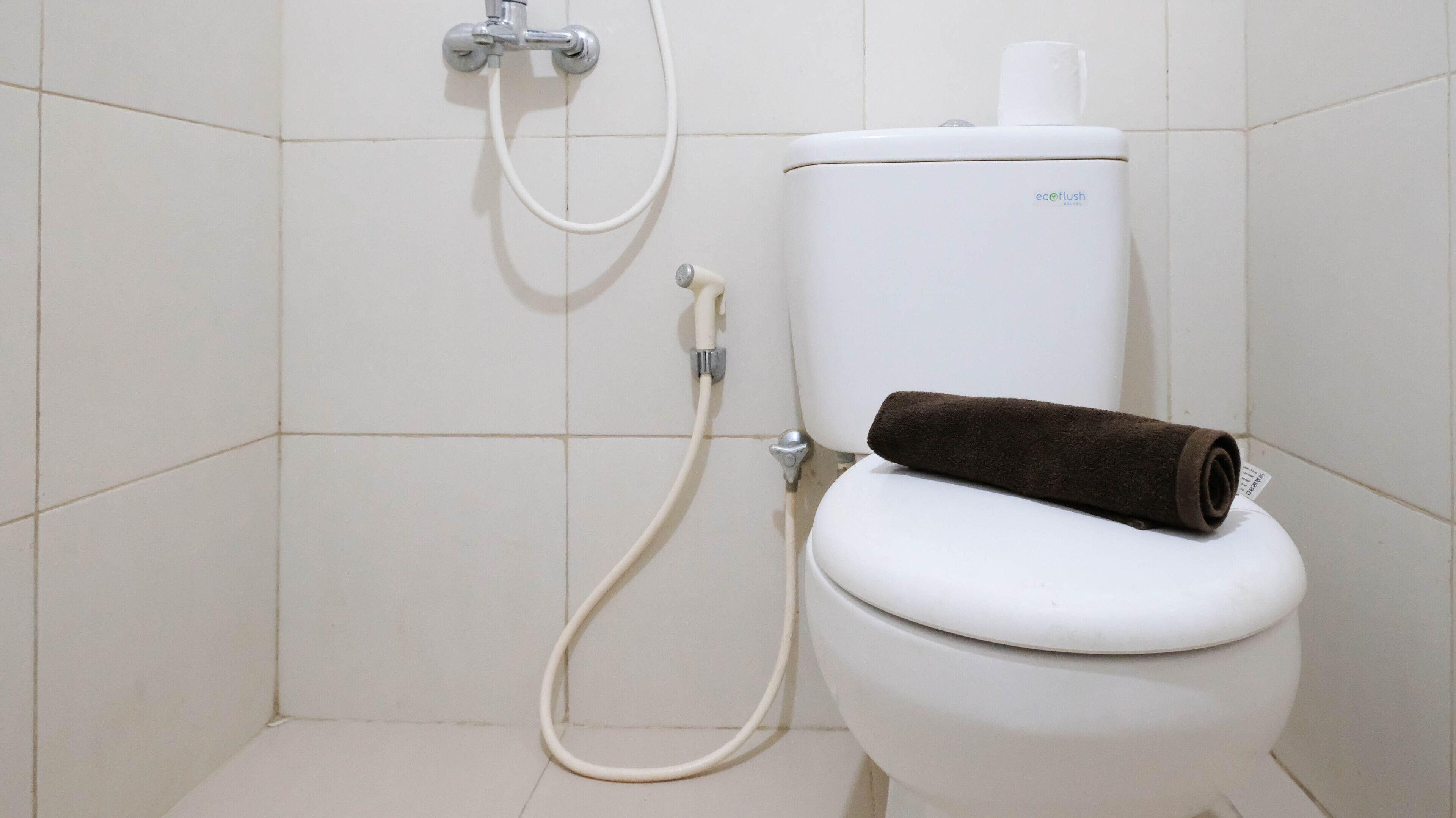 Eine weiße Toilette mit einem braunen Handtuch darauf, einem Toilettenpapierhalter und einem Duschschlauch.