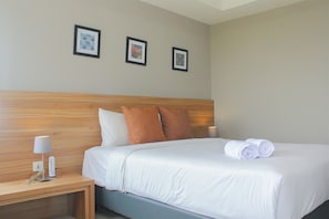 A neatly made bed with white linens, two brown pillows, and rolled white towels on a wooden headboard. There is a bedside table with a lamp and a small clock. Three framed pictures are hung on the wall above the bed. - Wonderful 1Br Apartment At Mustika Golf Residence (Cikarang)