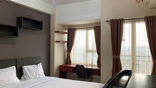 Ein Hotelzimmer mit Bett, Schreibtisch und Stuhl. Vor den Fenstern hängen Vorhänge und über dem Bett befindet sich ein Regal.
