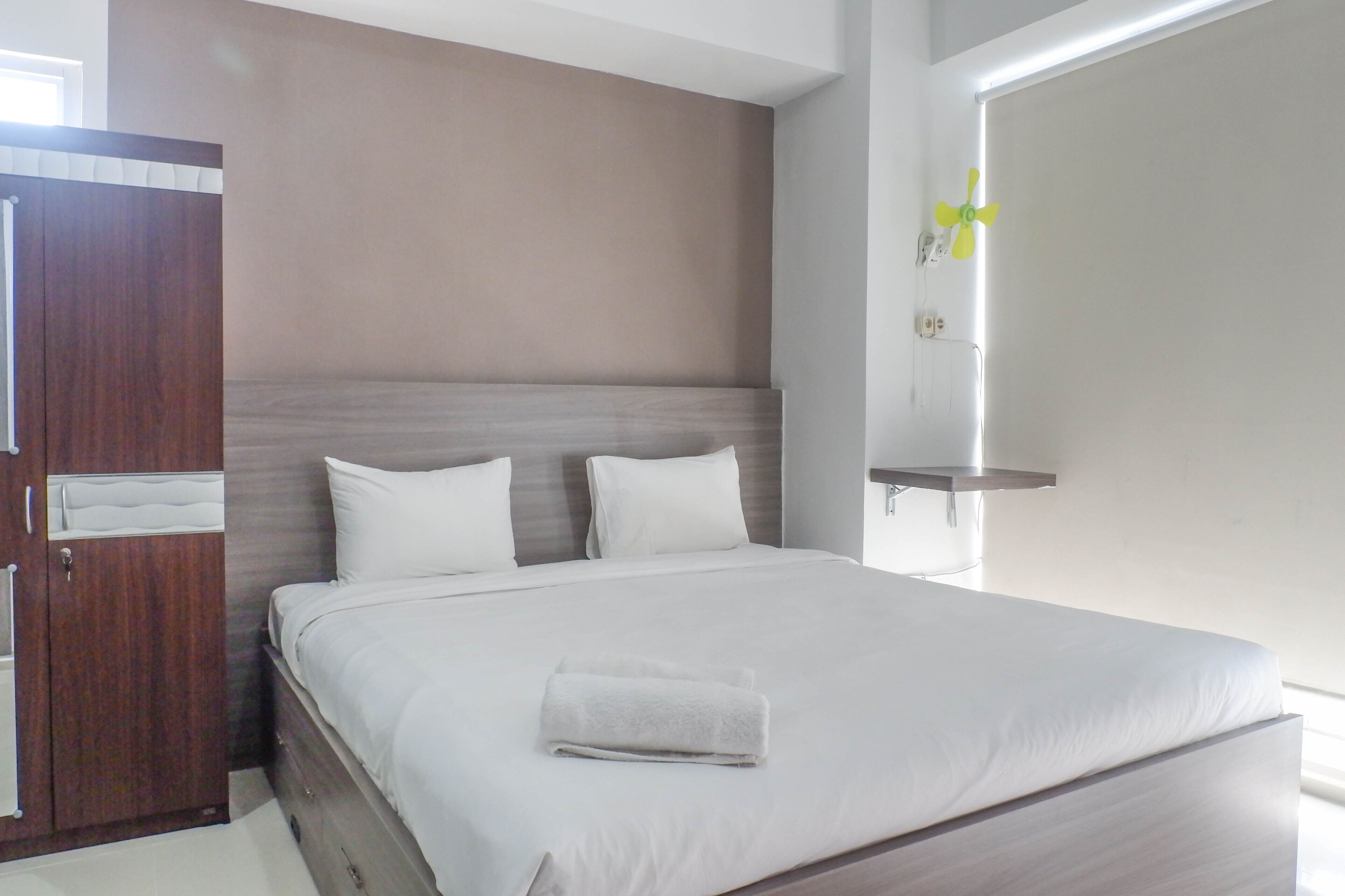 A neatly made bed with white linens and a folded towel, a wooden wardrobe, and a wall-mounted shelf with a fan.