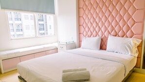 A neatly made bed with white linens and a rolled towel, a pink tufted headboard, a bedside table with a glass top, and a window with a view of buildings. - Warm And Comfort Stay 1Br Apartment At Silkwood Residences (Tangerang)