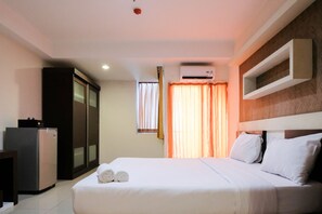 A hotel room with a bed, a desk, a refrigerator, and an air conditioning unit. - Cozy And Homey Studio Apartment At High Point Serviced (Surabaya)