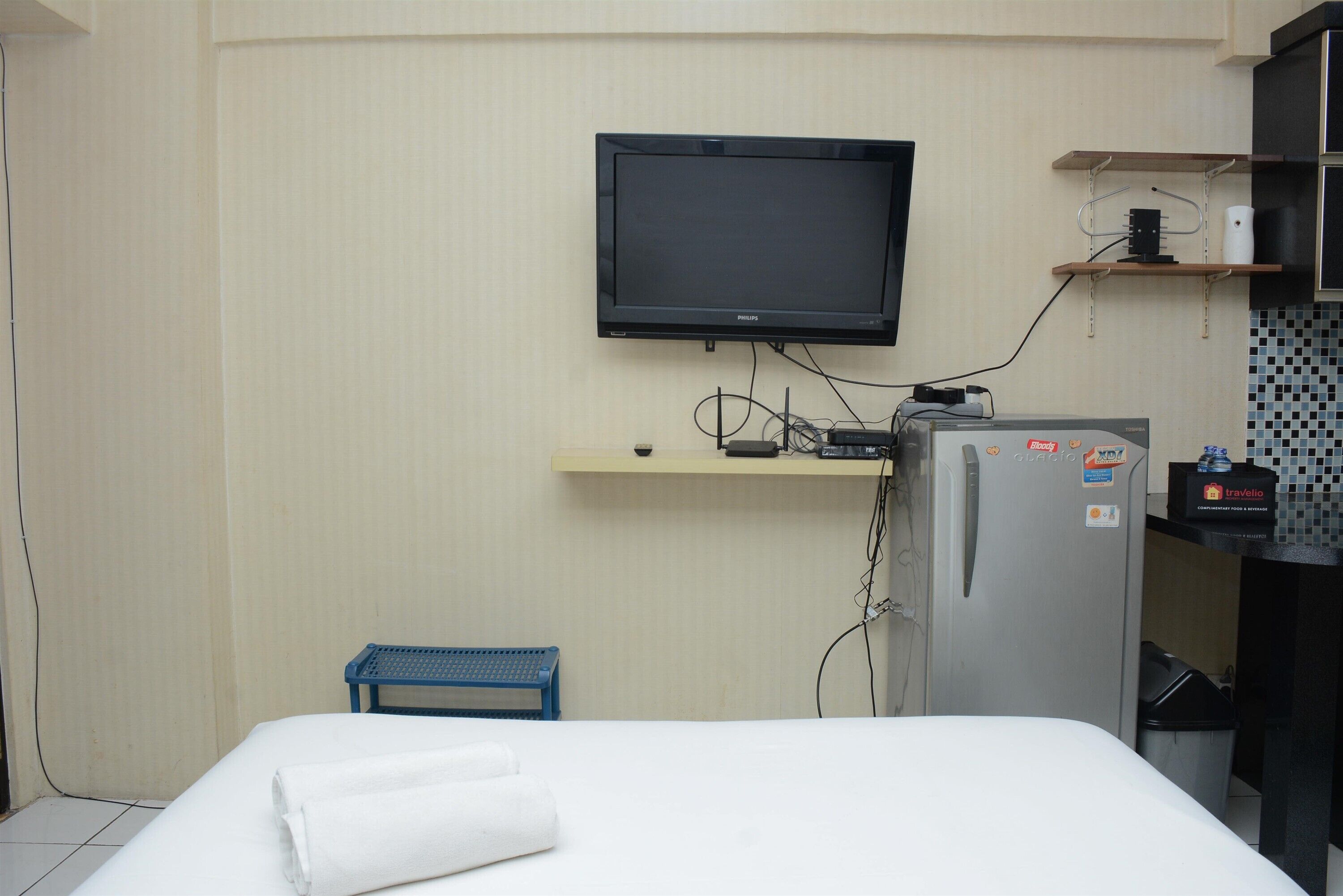 A hotel room with a bed, a television mounted on the wall, a small blue stool, a refrigerator, and a shelf with toiletries.