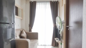 A living room with a sofa, a standing fan, and a door. - Comfort And Warm 2Br At Parkland Avenue Apartment (South Tangerang)