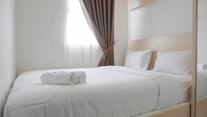 A neatly made bed with white linens and a rolled towel, a wooden headboard, and a window with sheer curtains. - Comfort And Warm 2Br At Parkland Avenue Apartment (South Tangerang)