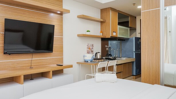 A modern hotel room with a flat-screen TV mounted on the wall, a wooden desk with a chair, a small kitchen area with a refrigerator and sink, and a bed with a white comforter. - Relax And Simply Studio Apartment At Parkland Avenue (South Tangerang)
