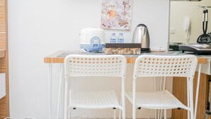 A small kitchenette with a coffee maker, water dispenser, and a kettle. There are two white chairs and a wooden table. A painting with coffee-related imagery hangs on the wall. - Relax And Simply Studio Apartment At Parkland Avenue (South Tangerang)