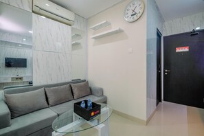 Uma sala de estar moderna com um sofá cinzento, uma mesa de centro de vidro com garrafas de água, um relógio fixado na parede e uma porta preta com uma placa vermelha.