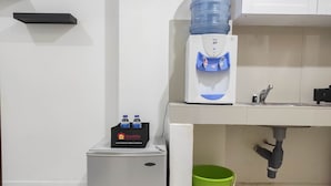 Une petite cuisine équipée d’un distributeur d’eau, d’un petit réfrigérateur, d’un seau vert et d’une étagère avec des bouteilles d’eau.