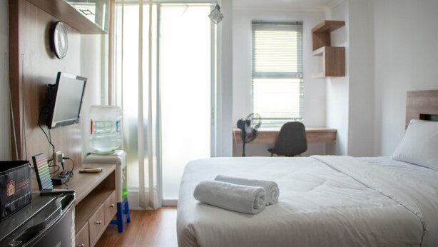 A hotel room with a bed, a desk, a chair, a water dispenser, and a television on a dresser. - Nice And Comfort Studio Apartment At Belmont Residence Puri (Jakarta)