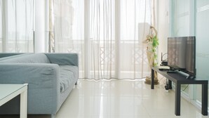A modern living room with a grey sofa, a TV on a black stand, and sheer curtains. - Strategic And Homey 2Br At Puri Garden Apartment Near Mall (Jakarta)