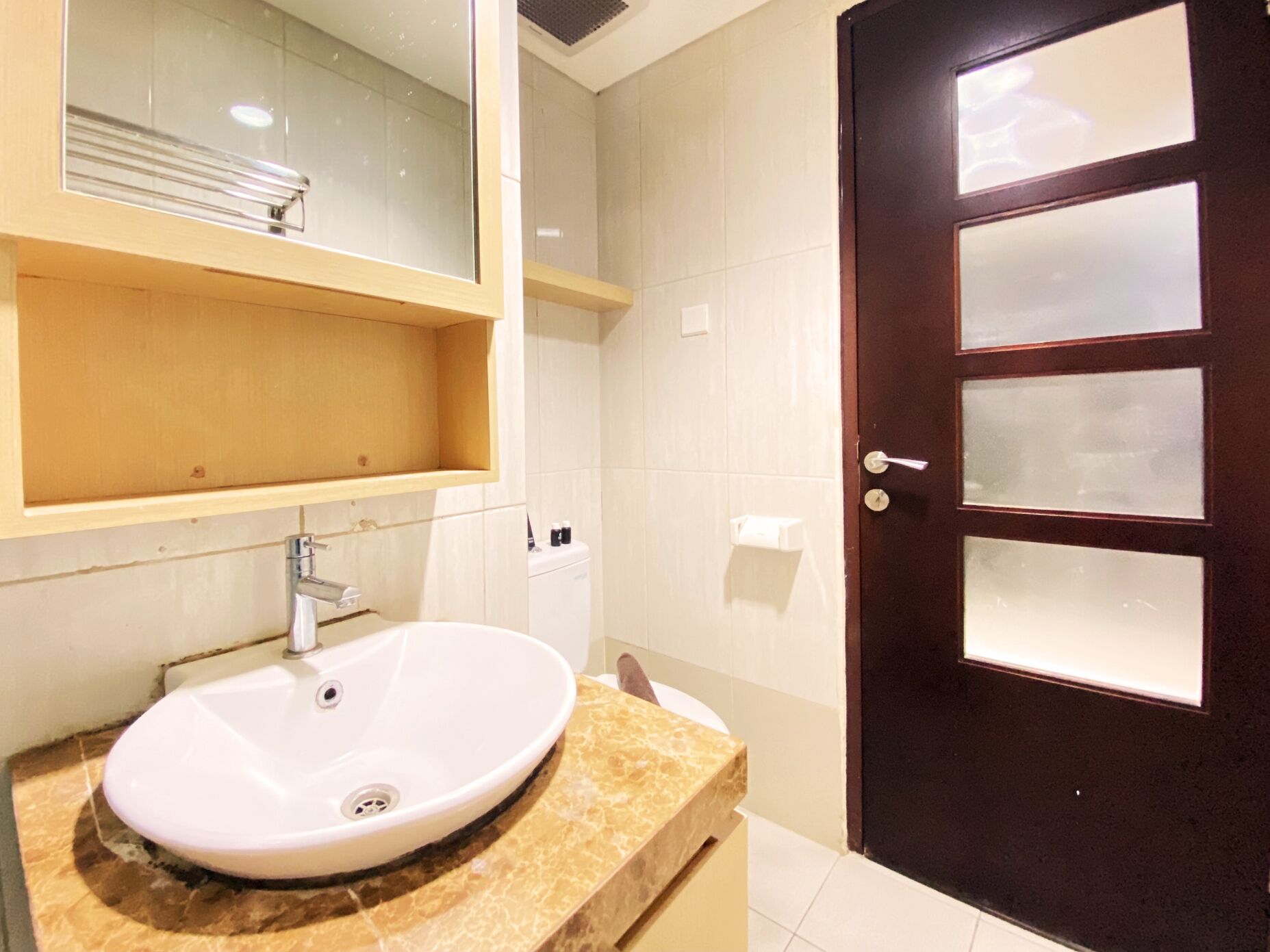 Un baño con un lavamanos blanco, un mueble de tocador de madera, un espejo y una puerta de madera oscura con paneles de vidrio esmerilado.