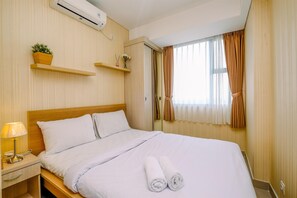 Uma cama bem arrumada com lençóis brancos e duas toalhas enroladas, uma mesa de cabeceira com abajur e telefone, e uma janela com cortinas.
