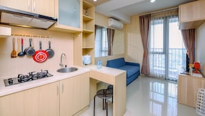A modern kitchen with a gas stove, sink, and hanging utensils. There is a blue sofa and a small table with a chair in the living area. - 1Br Apartment With Cozy Design At Royal Olive Residence (Jakarta)