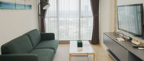 A modern living room with a green sofa, a coffee table, a TV on a stand, and a view of the city through large windows.