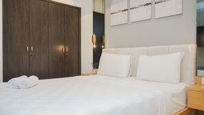 Ein ordentlich bezogenes Bett mit weißen Leinen und Kissen, ein Nachttisch aus Holz mit einer Lampe und ein dunkles Holzschrank.