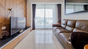 A modern living room with a flat-screen TV, wooden panel walls, a leather sofa, and a large window with sheer curtains. - Great Deal And Cozy 2Br At M-Town Signature Apartment (Gading Serpong)