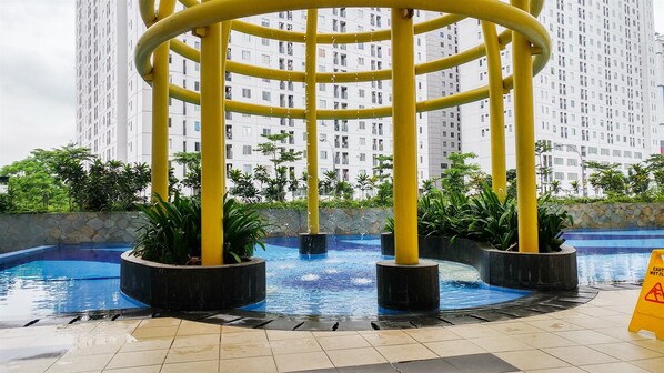 Une piscine dotée d’un élément décoratif aquatique circulaire et de piliers jaunes, entourée de verdure et avec des immeubles d’habitation en arrière-plan.