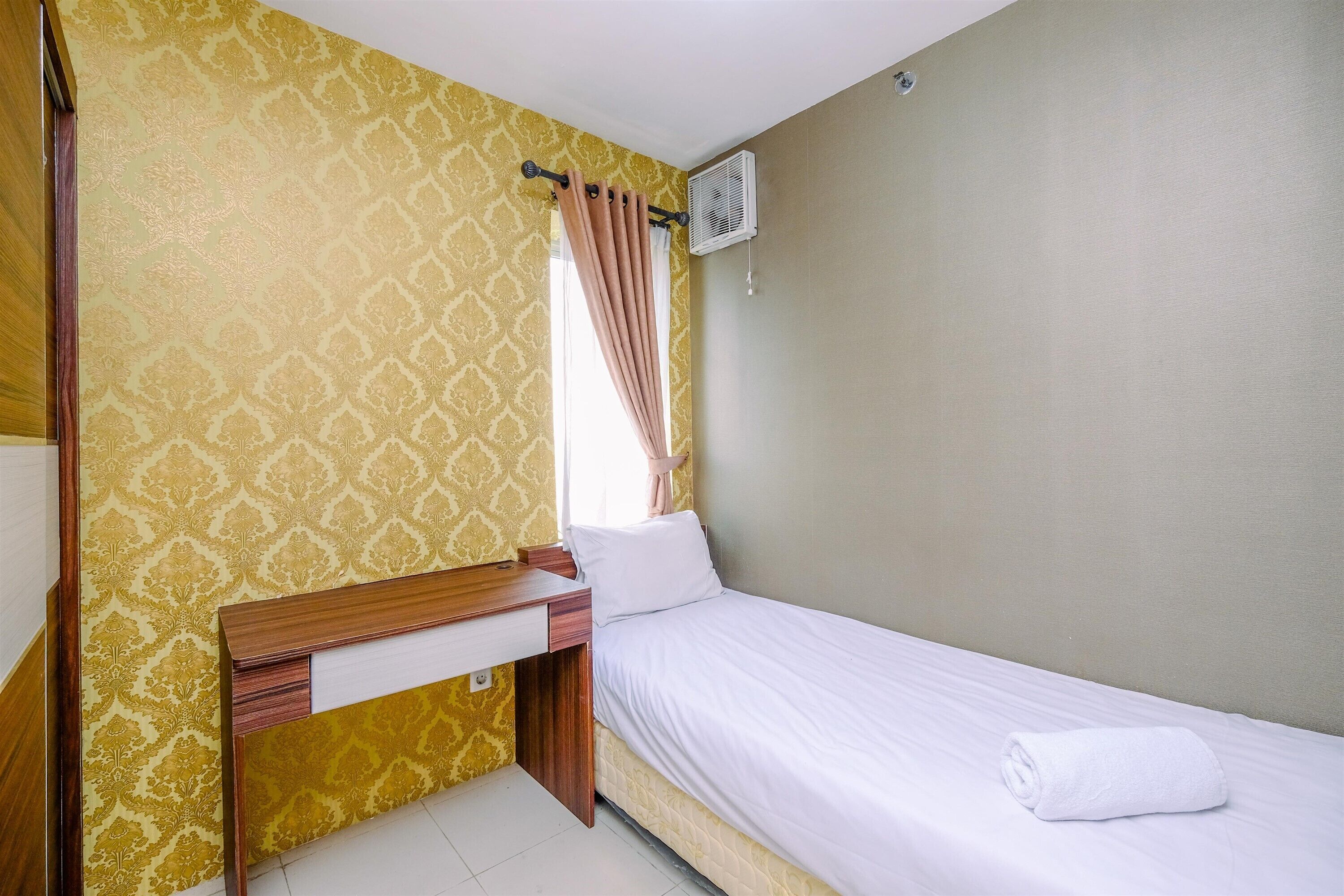 Una cama individual con sábana blanca y una toalla enrollada, una mesita de noche de madera, una ventana con cortina y un equipo de aire acondicionado en la pared.