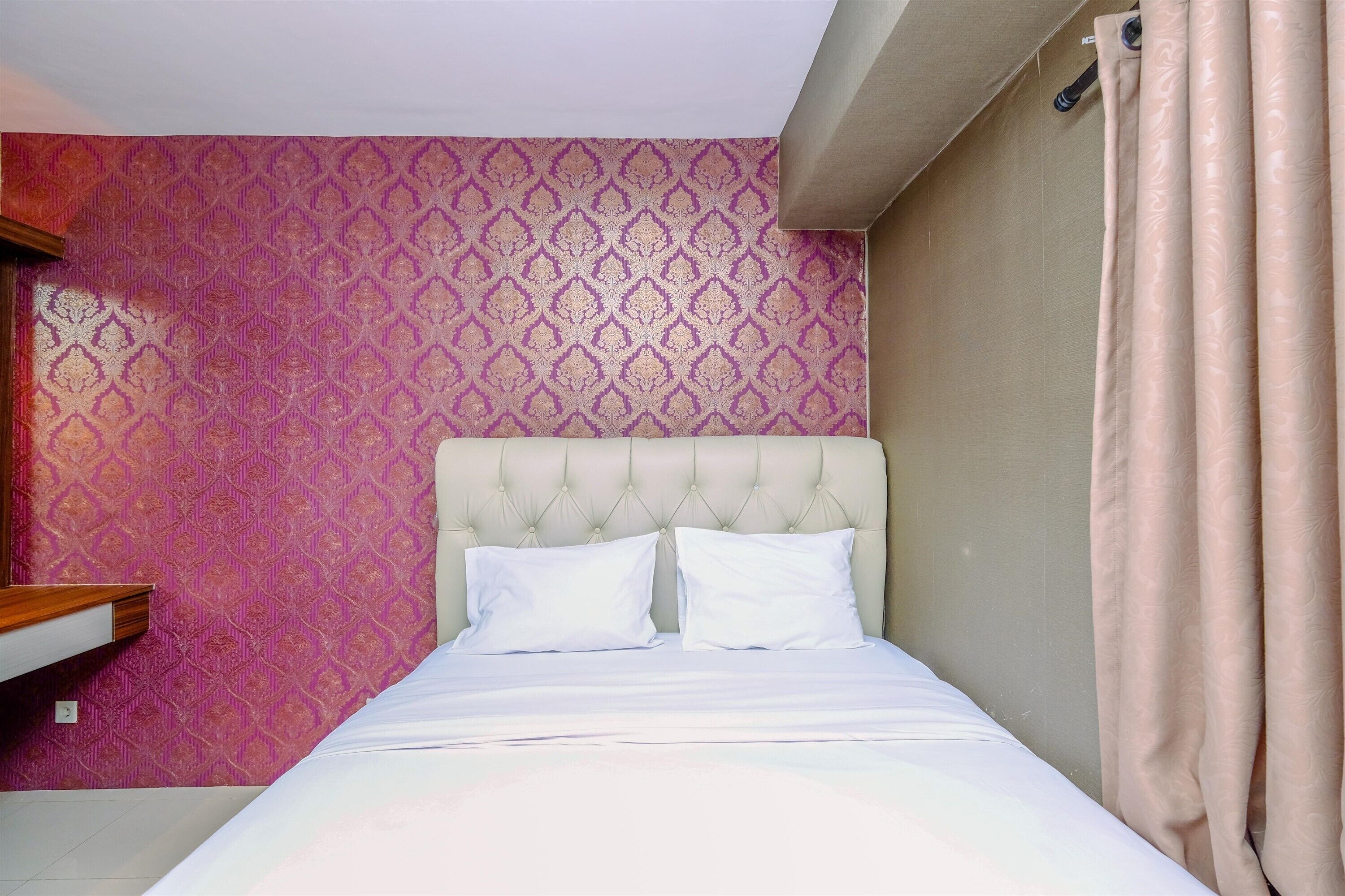 Een bed met een gebutste hoofdbord, twee witte kussens, en een effen muur met behang.