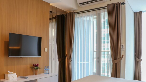 A hotel room with a flat-screen TV mounted on the wall, a bedside table with a tissue box and a potted plant, a bed with rolled towels, and a window with curtains. - Elegant And Comfy Studio At Tamansari Iswara Apartment (Bekasi)