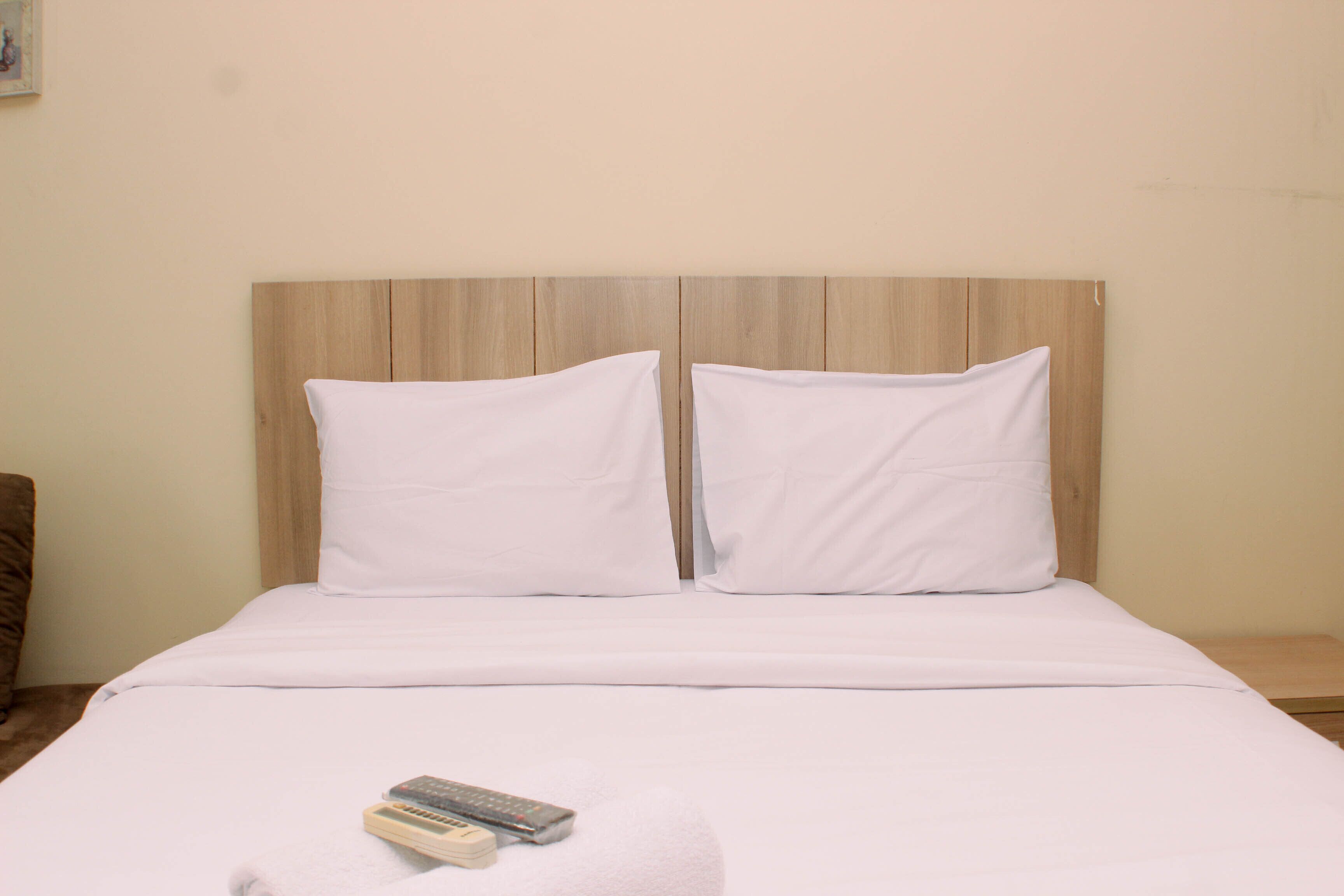 A neatly made bed with white bedding and pillows, a remote control, and a small nightstand with a lamp.