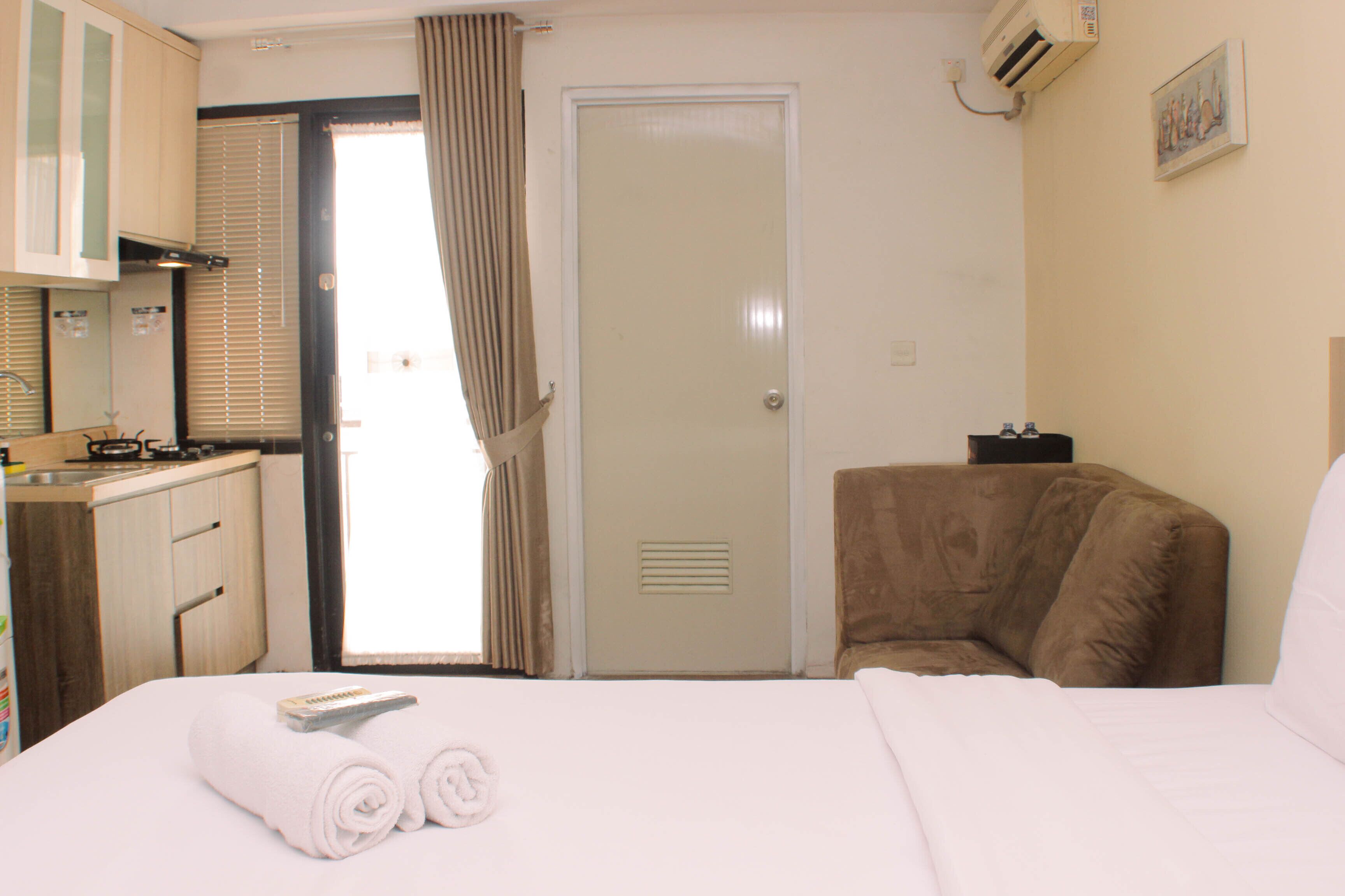 A hotel room with a bed, a small desk with a kettle, a chair, and a door with a window.