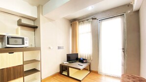 A compact living space with a wooden floor, a microwave on a shelf, a small table with a TV, and a sliding glass door. - Homey 1Br At Jarrdin Cihampelas Apartment (Bandung)