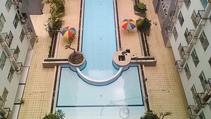 A swimming pool surrounded by a tiled walkway, with multiple umbrellas and potted plants. - Homey 1Br At Jarrdin Cihampelas Apartment (Bandung)
