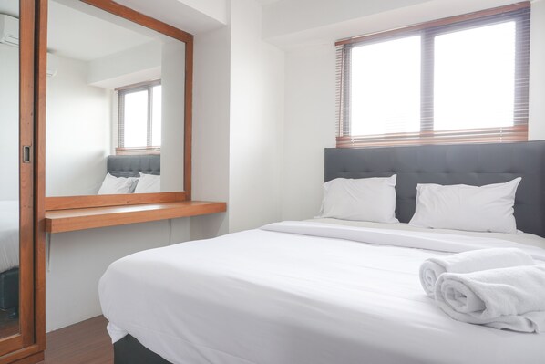 Un lit bien fait, avec du linge de lit blanc et une serviette enroulée, une table de chevet en bois et un grand miroir.