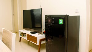 Une pièce avec un téléviseur à écran plat posé sur un meuble blanc, un réfrigérateur noir et une chaise en bois.