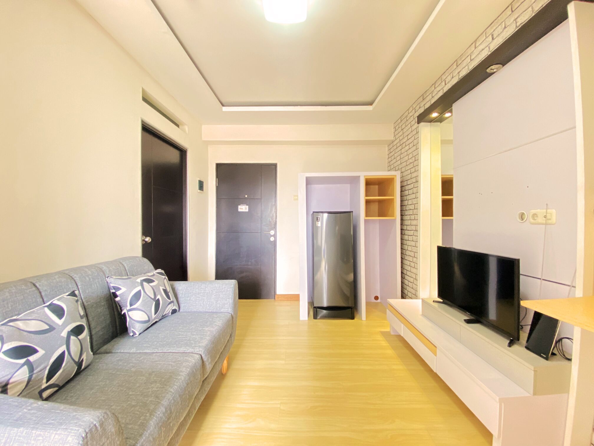 Un soggiorno moderno con un divano grigio, una TV a schermo piatto, una libreria a incasso e un angolo cottura con frigorifero e microonde.