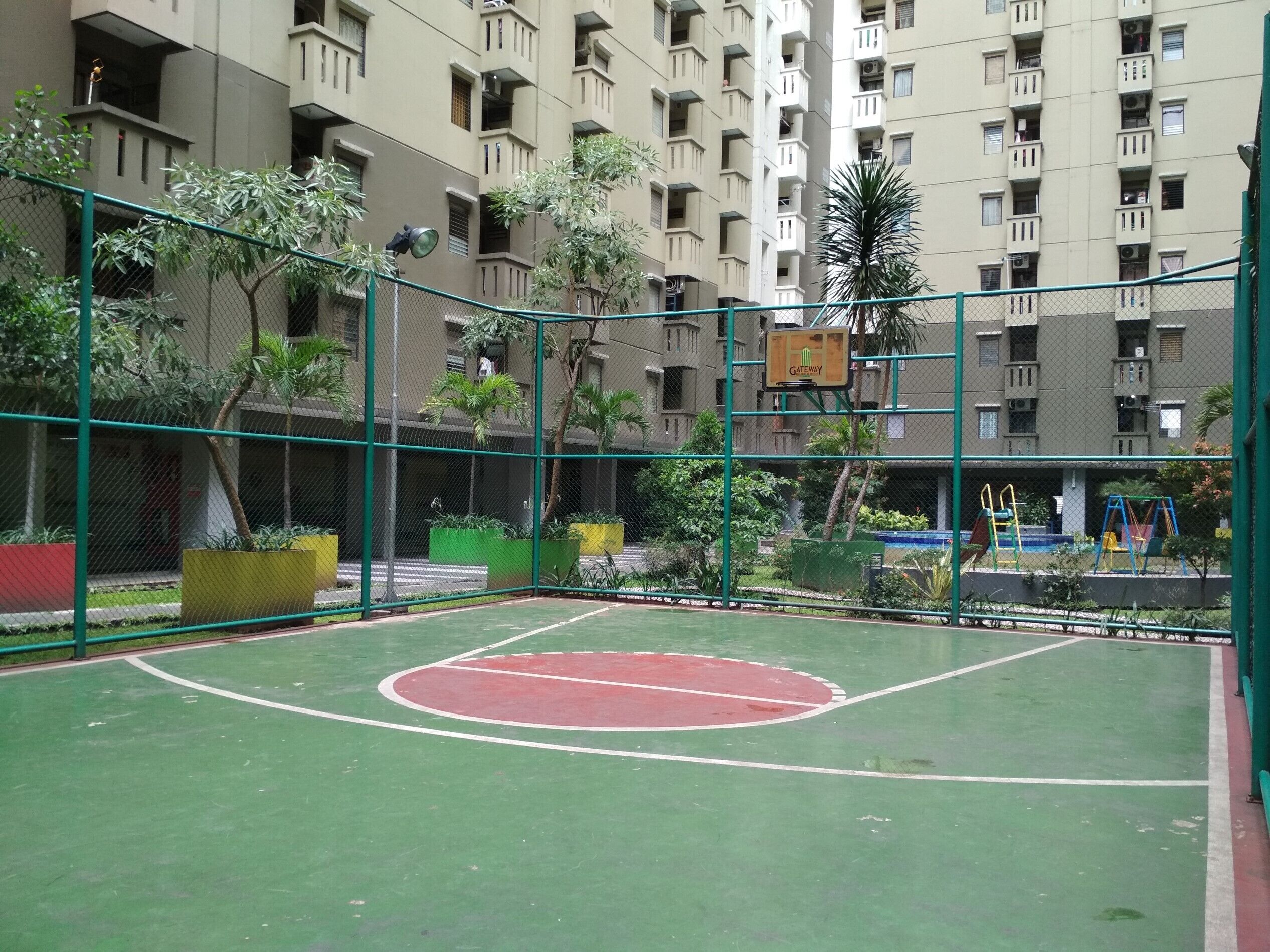 An outdoor basketball court surrounded by a green fence, with a playground in the background and a multi-story building with balconies.